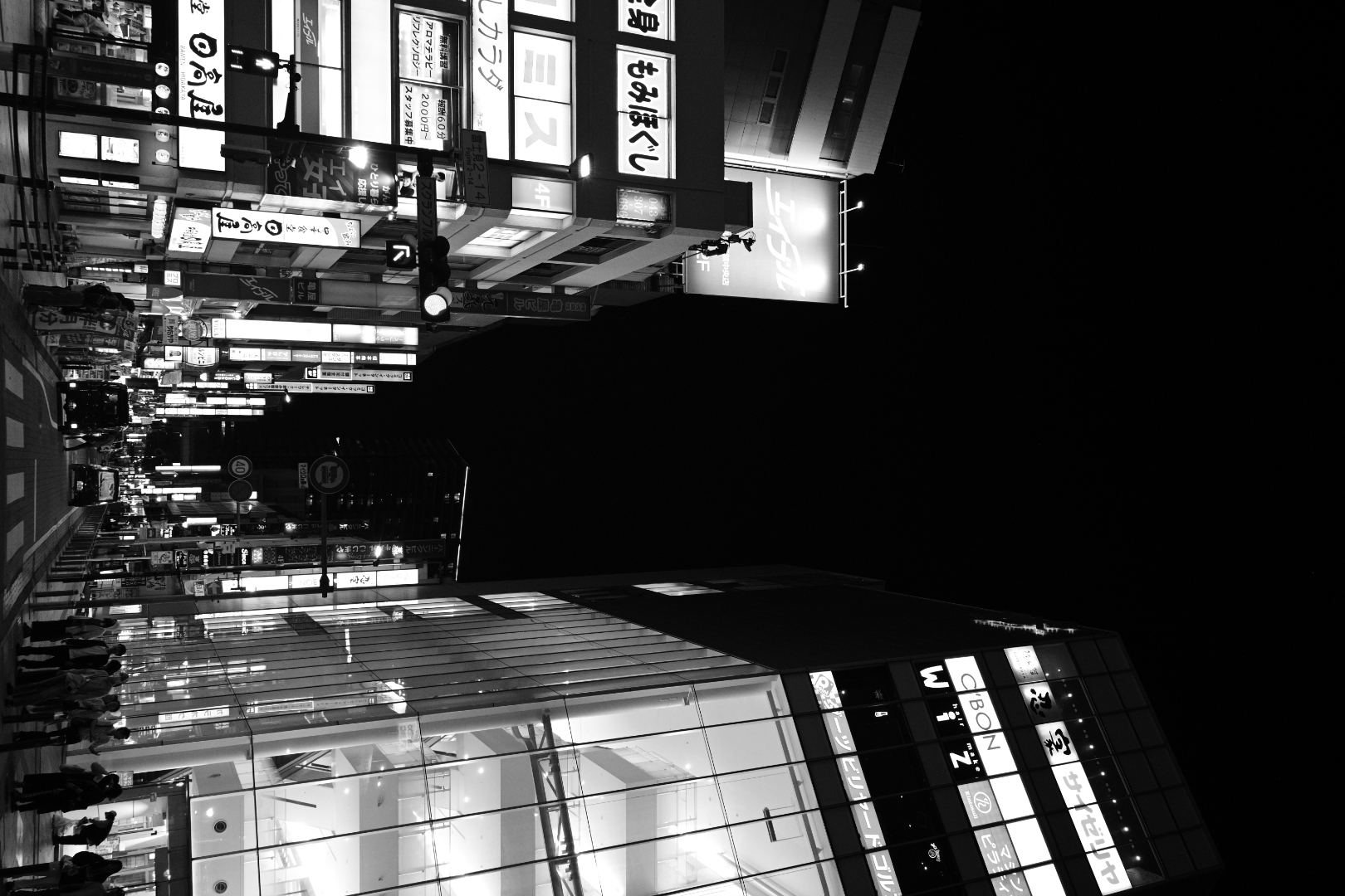 A city street at night in black and white, neon signs glowing between darkened buildings.